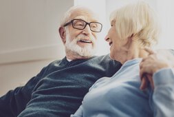 Ein Senior mit Brille sitzt neben einer Seniorin, umarmt sie und beide lächeln sich an. | © Fotolia 210582403 - Adobe Stock - Zinkevych